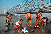 Kolkata, India - 28 January 2023: View of men bathing and washing clothes along the Hooghly River under the massive steel structure of the Howrah Bridge.