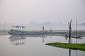 Bahmanbaria, Bangladesh - 30 January 2024: View of a tranquil river scene, where a weathered boat rests near a grassy bank, its reflection shimmering in the still water under a soft, hazy sky.