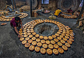 Bogura, Bangladesh - 03 May 2020: View of workers meticulously arranging fresh, golden-brown, circular food items around a central opening in a rustic, dimly lit industrial setting.