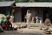 Chittagong, Bangladesh - 16 January 2019: View of a woman with walking stick stands near a tin house, while others work with terracotta pots in the yard.