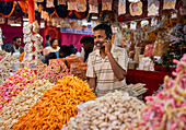Bogura, Bangladesh - 03 November 2023: View of a vibrant snack stall, overflowing with colorful sweets and savories, as a vendor engages in a phone conversation amid the bustling market scene.
