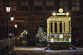 View of a brightly illuminated gate structure and Christmas tree sparkle against the dark stone buildings in Old Town, Gdansk, Poland.