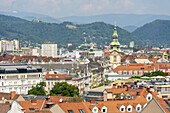 Blick auf in Terrakotta-Tönen leuchtende Dächer, die im Kontrast zu den gealterten Fassaden und den fernen, dunstigen Bergen stehen, während die Kirchtürme in die Höhe ragen, Graz, Steiermark, Österreich.