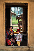 Dhangadhi, Nepal - 03. April 2023: Blick auf eine Mutter und ihre Kinder, eingerahmt von der strukturierten Tür eines traditionellen Lehmhauses, die die Wärme des Familienlebens unterstreicht.