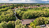 Aerial view of a river winding through dense green trees, crossing a road with moving cars, nestled near residential buildings, Guildford, Surrey, United Kingdom.