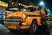 Kolkata, India - 28 January 2023: View of a bright yellow taxi cab reflecting the city lights, as a man peers inside, the iconic Blackberrys store sign glows above.