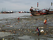 Ichanagar, Bangladesch - 03. August 2018: Blick auf spielende Kinder an einem schlammigen, mit Müll übersäten Ufer, während am fernen, grauen Horizont traditionelle Fischerboote liegen.