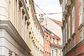 View of pastel buildings with ornate window frames and red tiled roofs under a soft, diffused sky, connected by overhead cables, Graz, Austria.