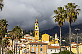 View of a vibrant yellow church tower rising above colorful buildings and palm trees against a dramatic sky, Menton, Provence-Alpes-Côte d'Azur, France.