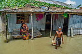 Bogura, Bangladesh - 07 March 2020: View of a mother with her child sitting outside a corrugated metal home as a boy walks through floodwaters.