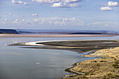 Das Wasser des Magadi-Sees ist in leuchtenden Rosatönen gehalten und steht im Kontrast zu der kargen Landschaft und den fernen Bergen unter einem weiten Himmel, Magadi, Kajiado, Kenia.