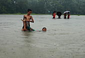 Mirsarai, Bangladesch - 01. Januar 2000: Blick auf zwei Jungen, die vergnügt auf einem überschwemmten Feld spielen, während in der Ferne Menschen mit Regenschirmen stehen und eine Szene der Resilienz schaffen.