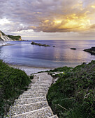 Blick auf Steintreppen, die zum Strand Man O War hinunterführen, unter einem leuchtenden Himmel, der sich in Orange- und Violetttönen auf dem ruhigen Meer spiegelt, Wareham, Dorset, England, Vereinigtes Königreich.