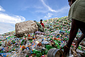 Chittagong, Bangladesh - 22 September 2022: View of a vast, colorful mountain of discarded plastic bottles under a bright sky, where a young boy and woman work amidst the waste.