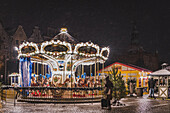 View of a brightly lit carousel stands amidst the snowy Old Town square, its golden lights casting a warm glow on the cobblestone, Gdansk, Poland.
