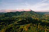 Luftaufnahme einer grünen Bergkette, die von einer weichen, ätherischen Wolke gekrönt wird, die einen Kontrast zu den tiefen Schatten und den sonnenbeschienenen Hängen bildet, San Enrique, Western Visayas, Philippinen.