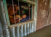 Bogura, Bangladesch - 07. März 2020: Blick auf ein Kind, das auf einem Kissen liegt und aus einem Fenster in einer von Überschwemmungen umgebenen Wellblechkonstruktion schaut.