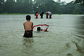 Mirsarai, Bangladesch - 01. Januar 2000: Blick auf Kinder, die durch die Fluten waten, Regenschirme, die in der Ferne inmitten einer düsteren, regennassen Landschaft wippen.