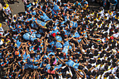 Mumbai, India - 19 August 2022: View of a human pyramid formed by young men, their blue and orange attire a striking contrast against the surrounding crowd's white garments.