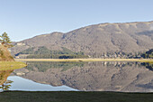 View of tranquil lake mirroring rolling hills under a clear sky, with a hint of autumnal hues and the soft embrace of nature's quiet beauty, Bagnoli, Provincia di Avellino, Italy.