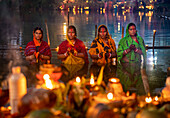 Bogura, Bangladesh - 20 November 2023: View of women in vibrant saris, amidst flickering candles and offerings, praying during a riverbank Chhath Puja celebration.