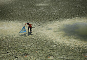 Mirsharai, Bangladesh - 17 August 2018: View of a lone boy with a net stands on a cracked, parched earth, the dry landscape contrasting with a small pool of stagnant water.