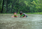 Mirsarai, Bangladesch - 01. Januar 2000: Blick auf zwei Kinder, die hüfthoch im Hochwasser stehen, ihre Gesichter leuchten im hellen Grün der Blätter, die sie vor dem grauen Himmel halten.