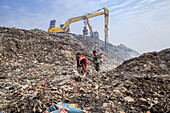 Chittagong, Bangladesh - 12 February 2024: View of children navigating the immense, stark landscape of a garbage dump, overlooked by towering yellow machinery amidst a hazy sky.