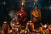 Bogura, Bangladesh - 20 November 2023: View of women holding small flames in the dark, their faces softly lit, amidst flickering earthen lamps and offerings on the water's edge.