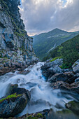 Blick auf Wasserfälle, die über moosbewachsene Felsen hinabstürzen, unter dem wachsamen Blick hoch aufragender Felsen und entfernter Gipfel, Vysoké Tatry, Region Prešov, Slowakei.