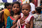 Bogura, Bangladesh - 03 November 2023: View of a tender moment as a young girl lovingly holds a baby, both adorned with traditional markings, amidst a bustling crowd near a temple.