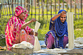 Bogura, Bangladesh - 30 May 2018: View of women sitting on the ground, their vibrant pink and blue clothing contrasting with the pale fibers they work with, surrounded by lush greenery.