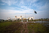 View of a golden kirpan statue gleams in the foreground, contrasting against the white gurdwara under a vast sky with the Pakistani flag waving, Kartarpur, Punjab, Pakistan.