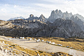 Blick auf Wohnmobile und Autos, die in einer kargen Landschaft vor dem Hintergrund der schroffen, grauen Gipfel der Drei Zinnen geparkt sind, Misurina, Belluno, Italien.