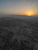 Aerial view of the sun setting over the rugged, mountainous terrain, casting long shadows and highlighting the stark beauty of the landscape, Aaqoura, Mount Lebanon Governorate, Lebanon.