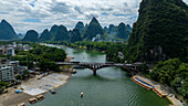 Aerial view of the Li River flowing beneath a bridge, surrounded by towering karst mountains against a cloudy sky, Yangshuo, China.