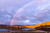 View of a vibrant rainbow arcs gracefully over the tranquil lake, illuminating the golden cliffs and rugged landscape, Lyle, Washington, United States.
