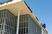 View of a modern building with glass facade and rooftop greenery under a clear sky, flags waving gently in Athina, Attica, Greece.