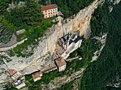 Aerial view of a church clinging to a sheer cliff face, a testament to faith and architecture, against a backdrop of lush greenery, Madonna della Corona, Veneto, Italy.