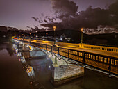 Aerial view of the Cavendish bridge illuminated by warm yellow lights stretching across the water under a moody sky, Mahebourg, Grand Port District, Mauritius.