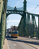 View of a yellow tram traversing the green iron framework of a bridge under a bright sky, casting shadows on the road, Budapest, Hungary.