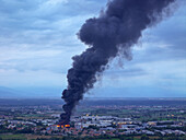 Pianezza, Italy - 04 September 2025: Aerial view of a dense column of black smoke rising ominously from the Euro Stamp plant, stark against the muted tones of the surrounding urban landscape.