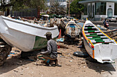 Dakar, Senegal - 12 May 2025: View of a man crouching near fishing boats on the sandy Soumbédioune beach, under the harsh sun, painting the hull of a boat.