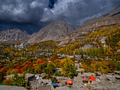 Blick auf dramatische Berge, die sich über einem Dorf erheben, das in den Farben des Herbstes leuchtet, ein Wandteppich aus Rot, Orange und Gold unter einem düsteren Himmel, Hunza Nagar, Gilgit Baltistan, Pakistan.
