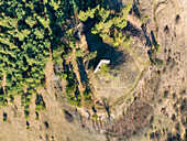 Aerial view of a lone structure atop a small hill surrounded by a ring of dry grass and contrasted by dense green trees, Podzámcok, Banská Bystrica Region, Slovakia.