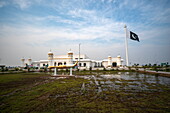 View of shimmering pools reflecting the white-washed architecture and fluttering Pakistani flag under a vast sky, Kartarpur, Punjab, Pakistan.