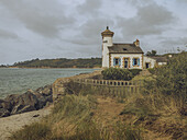 View of the quaint The Phare de Nantouar with blue shutters perched on a rocky shore where the turbulent sea meets the land, Louannec, Bretagne, France.