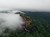 Aerial view of the ancient rock fortress of Lion Rock piercing through the swirling mists, a symphony of green jungle against the grey skies, Sigiriya, Central Province, Sri Lanka.