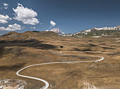 Luftaufnahme einer kurvenreichen Straße, die sich durch die goldene, sonnenüberflutete Hochebene in Richtung des majestätischen Gran Sasso-Gebirges unter einem strahlend blauen Himmel schlängelt, Campo Imperatore, Abruzzen, Italien.