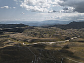 Luftaufnahme der kurvenreichen Straßen, die sich durch die zerklüftete Landschaft von Campo Imperatore schlängeln, wo die Schatten über das sonnenbeschienene Gelände tanzen, Gran Sasso, Abruzzen, Italien.